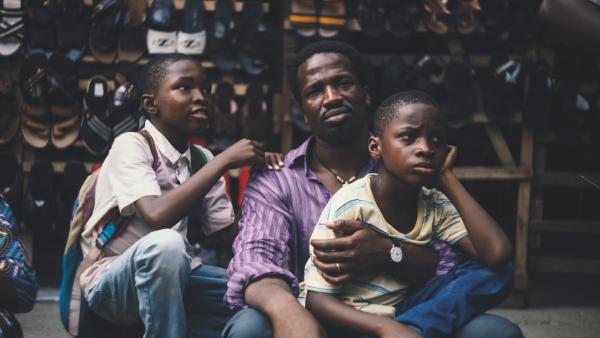 A man sits with two children resting against him while another child sits nearby. They are seated outdoors in front of a display of shoes arranged on shelves.