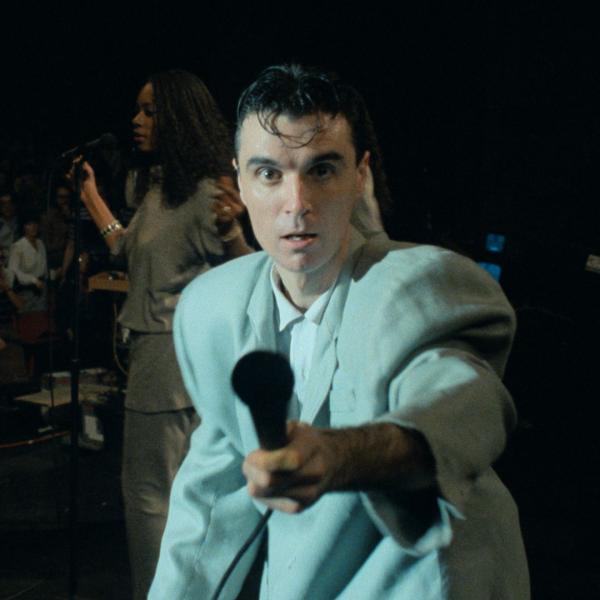 On stage in an oversized grey suit, David Byrne holds up a microphone to the camera. A singer and crowd are visible in the background.