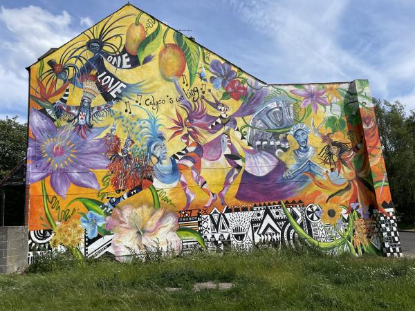 mage of a large mural on the  very colourful mural painted on the gable end of a row of terraced  post war houses. at the bottom of the muralis ovr grown grass on uneven ground.