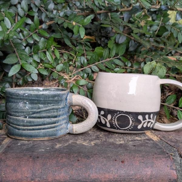 A photograph of two handmade mugs, one is blue and the other is white with black decoration