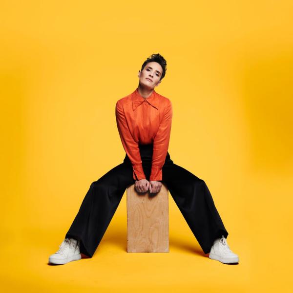 Jen Brister sits on a wooden block with her head tilted to the side, wearing long back trousers, white trainers a bright orange shirt. 