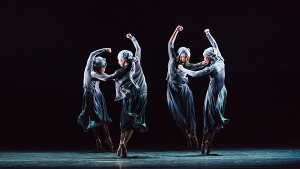 Four performers dance on stage in pairs. They have their arms in the air. 