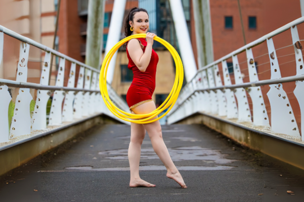 A woman in a red leotard stands with a yellow hula hoop on a bridge