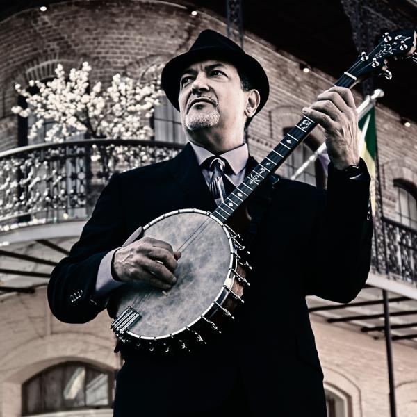 Shot from below, Don Vappie stands at a street corner, playing a banjo and looking into the distance, wearing a black suit and pork pie hat.