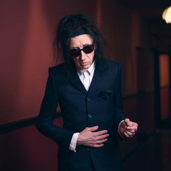 Dr John Cooper Clarke casually poses in a black suit, wearing his iconic sunglasses.
