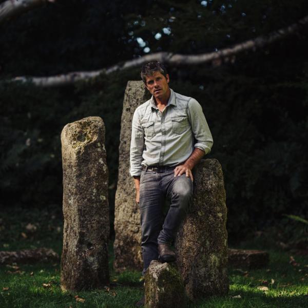 Seth Lakeman leans against a mossy standing stone in a woodland setting.