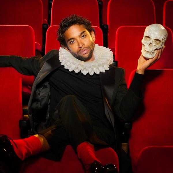 Hiran Abeysekera as Hamlet sits in contemporary red theatre seats, casually tilting his head to the side and holding a skull in his hand.