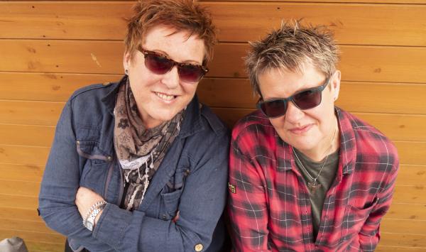 Picture of two women with short scruffy hair and cheeky smiles, they are wearing casual clothes and sunglasses, they look like their gig will be a lot of fun