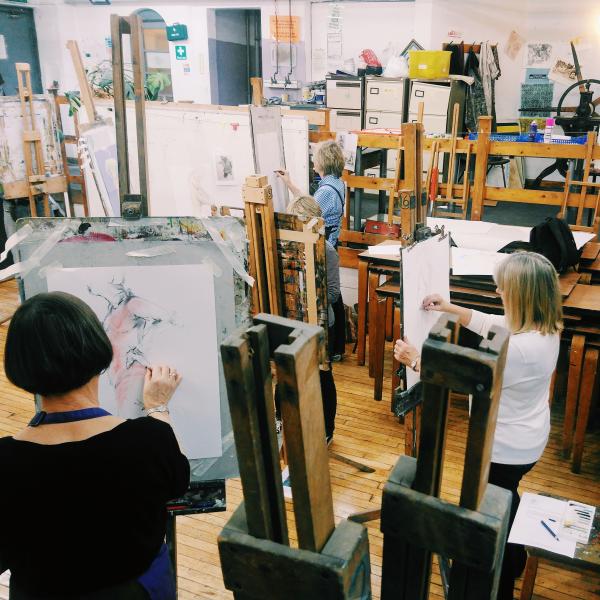 A photo of 3 women stood up at easels, they are all drawing a life model. the woman in the foreground is using a pink pencil to add colour to her work