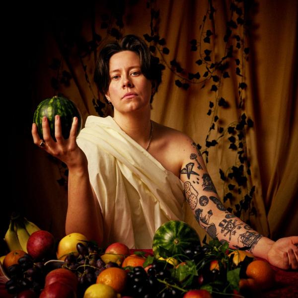 Chloe Petts in a toga sat in front of a table of fruit holding a watermelon.