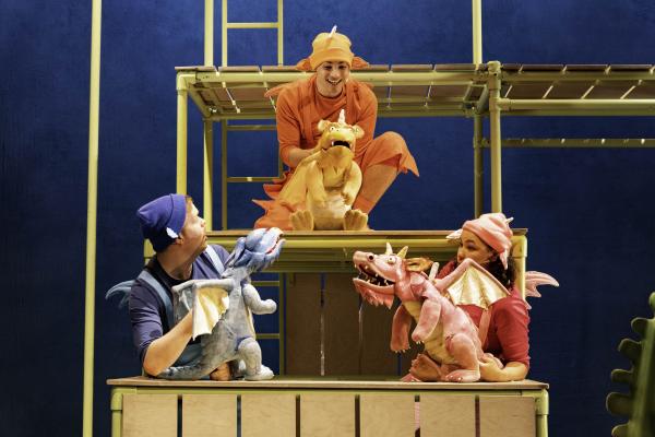 Three actors control puppets of different dinosaurs, one is pink, one is blue and one is orange.