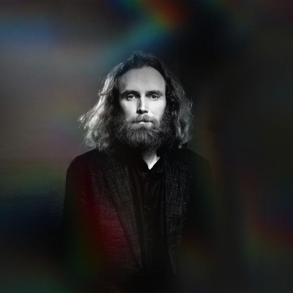 In black and white, Rob Auton with dark shoulder-length hair and beard dressed in a black suit stands alone and serious against an empty backdrop.
