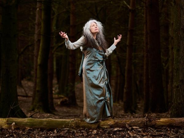 A woman in silk robes stands in a wood with arms in the air