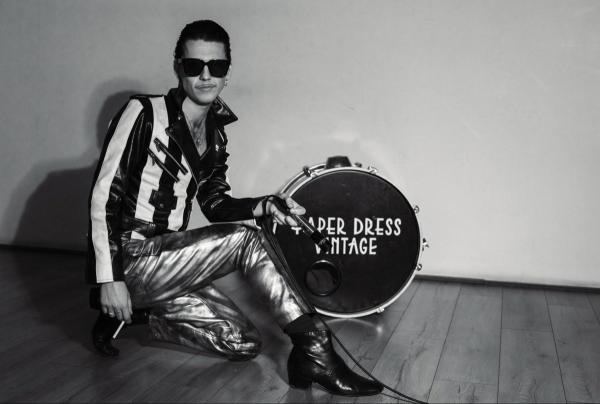 A black and white picture Tiss, wearing dark sunglasses and shiny trousers. Posing with a cigarette and drum.