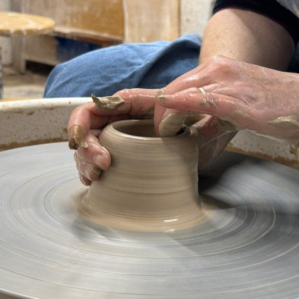A photograph of hands working a clay vessel whilst is spins on a pottery wheel