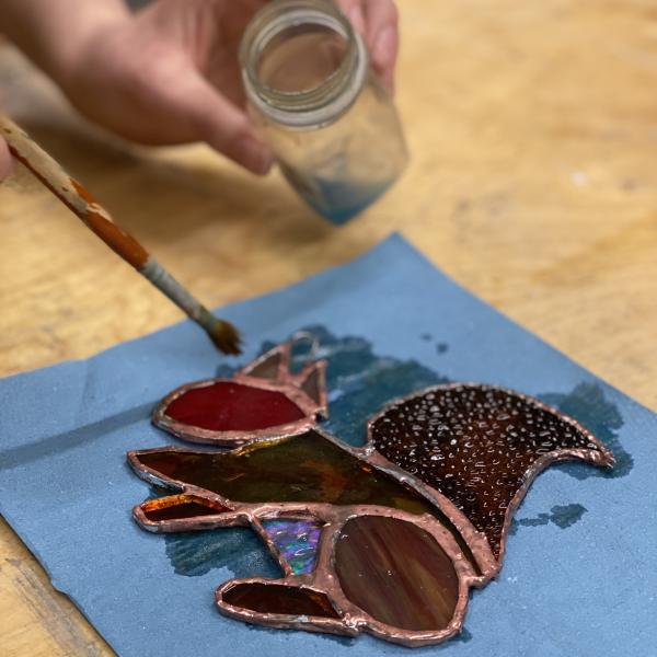 A photograph of a stained glass squirrel on some blue paper with a hand poised over it with a paintbrush  