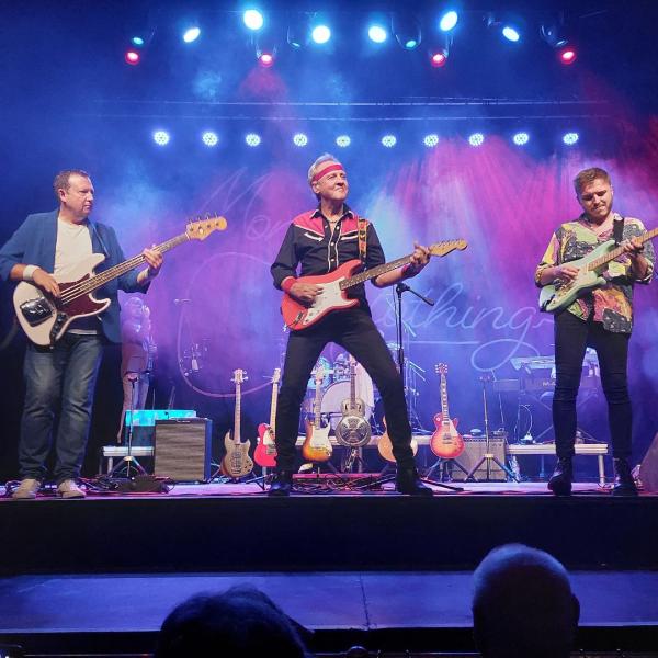 Three guitars perform on stage, standing in a row, each playing electric guitars. 