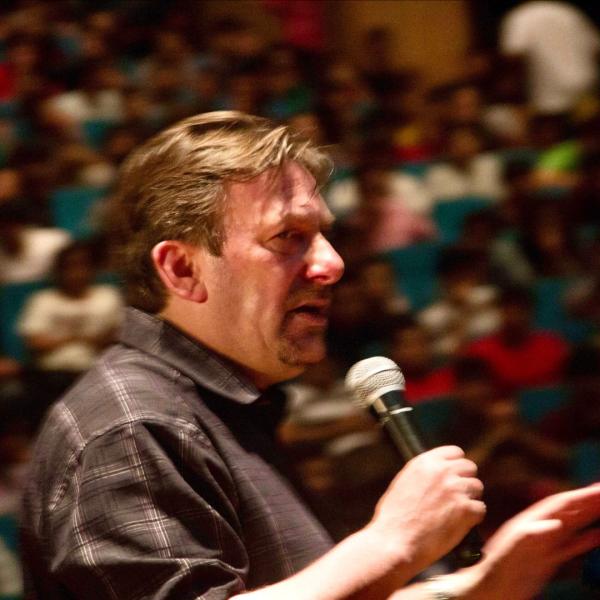 Andrew Newton performs to a packed blurry crowd, speaking into a handheld microphone and wearing a black shirt.