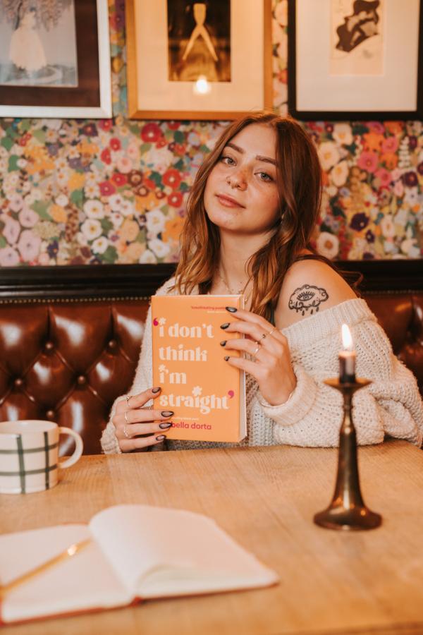 A woman with a cloud tattoo on her shoulder sits at a wooden table, holding the book 'how to leave your psychopath' by Pura. She is in a cozy room with floral wallpaper and a lit candle.
