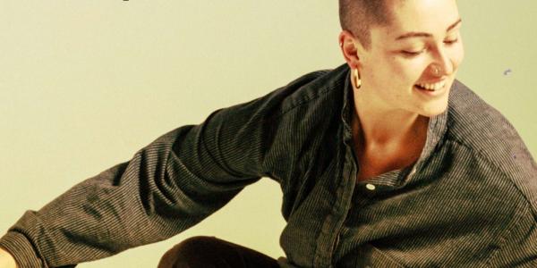 Image of person with make up and a shaved head. Image is focused on the head, arm and shoulders. They are smiling and look to be in the middle of a dance