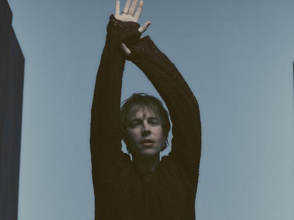 Tom Odell standing with arms raised above their head against a plain background, wearing a dark long‑sleeved top.