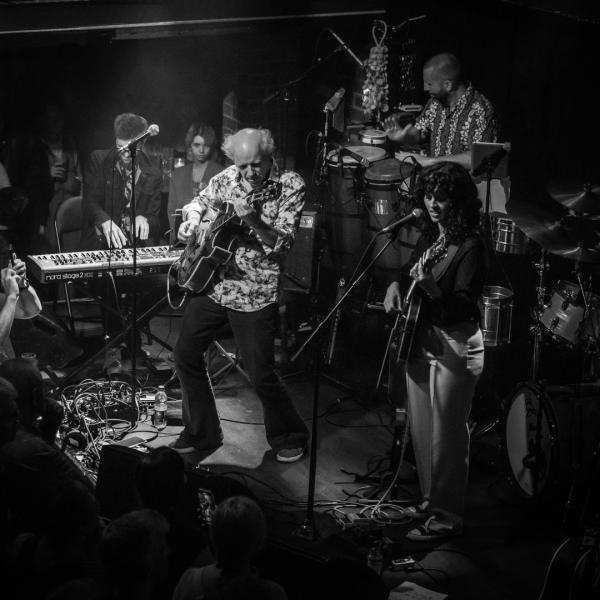 The band Hejira perform in a crowded stage setting. From left to right, there is a keyboard player, guitarist, drummer and guitarist-vocalist beneath the moody spotlight.