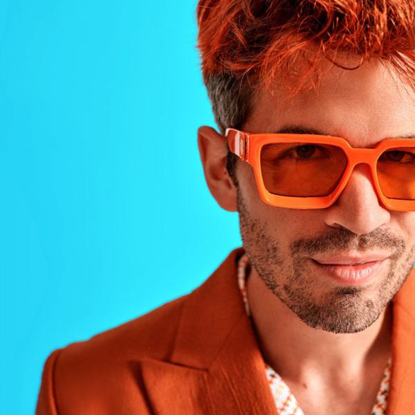 Ivo Graham smirks at the camera, in a bright orange suit and sunglasses, with orange-dyed hair.