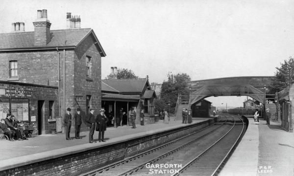 Historic black-and-white photograph of Garforth railway station. Brick station buildings line the left platform, where several people stand and sit beneath railway posters. Curving railway tracks run through the centre of the scene beneath a covered footbridge linking the platforms. Text at the bottom of the photograph reads “GARFORTH STATION”.