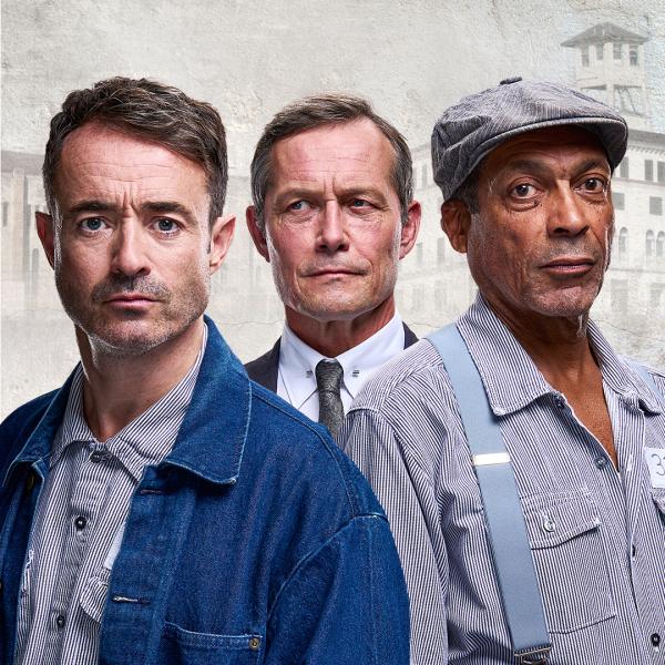Two men in vintage prison gear look sternly at the camera, accompanied by a suspicious suited man behind them. In the background is a stylised illustration of a prison block.