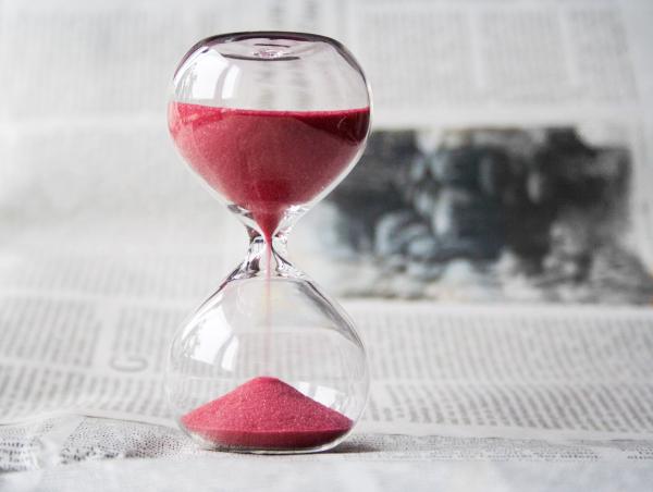 Close-up of a modern glass hourglass with pink sand against a blurred background of newspaper text, symbolizing the passage of time or a deadline.