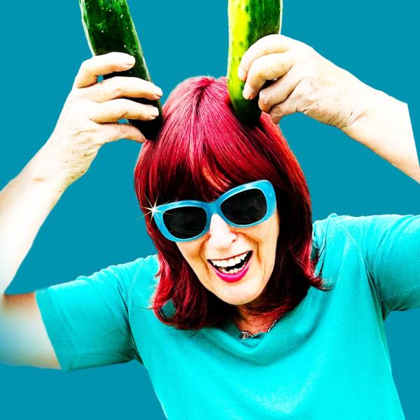 Janet Street Porter with bright red hair and blue sunglasses, laughing and holding up two cucumbers like horns above her head.