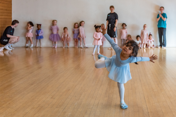 A small girl in a blue tutu balances on one leg smiling at the camera as other children look towards her
