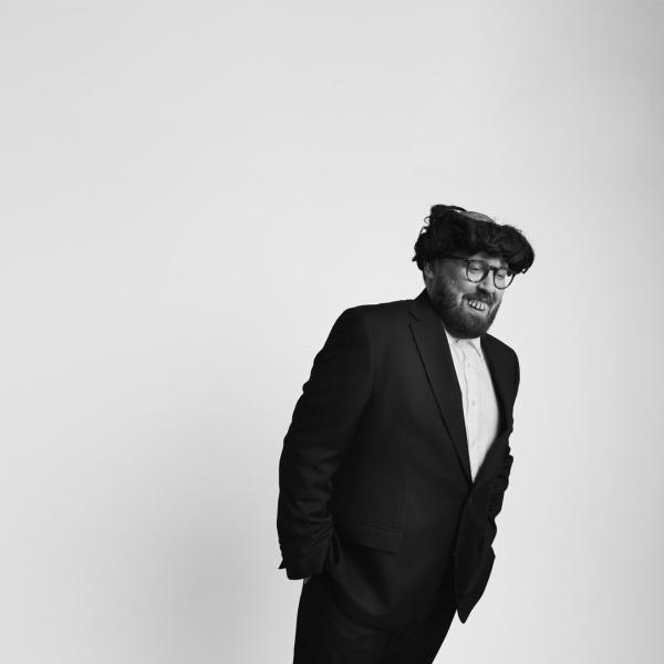 Black and white photo of John Kearns in a simple suit, wearing a comic wig and fake teeth. He tilts sideways against a blank backdrop.