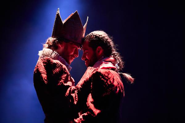 Two men are in an embrace, looking into each other's eyes. One of them has a large crown on, and there is a dark background.