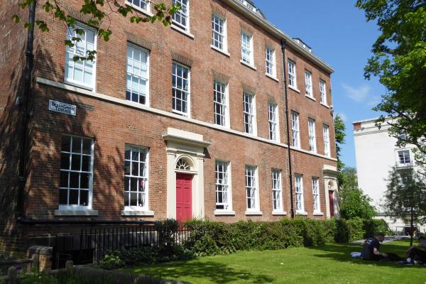 Three-storey red-brick Georgian terrace with white sash windows and red entrance doors, viewed from a grassed garden. Trees frame the building, and a street sign on the corner reads “Beech Grove Terrace”. Two people sit on the lawn at the right-hand side of the scene.