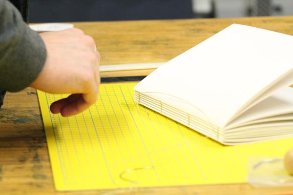 A hand reaching out to a coptic bound white notebook, which is positioned on a bright yellow cutting mat on a wooden table.
