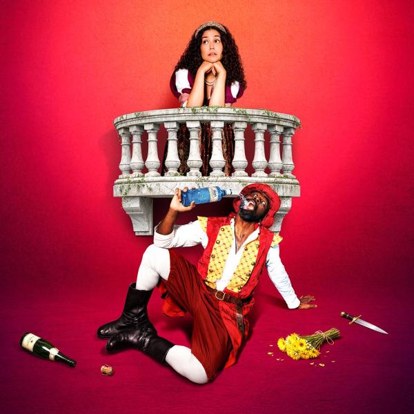 Stood at a prop balcony, an actress in period costume rests her head in her hands and looks off-camera. In front of her, another actor is sat on the floor in a red tunic and boots, drinking from a bottle of gin beside an empty bottle and discarded dagger.