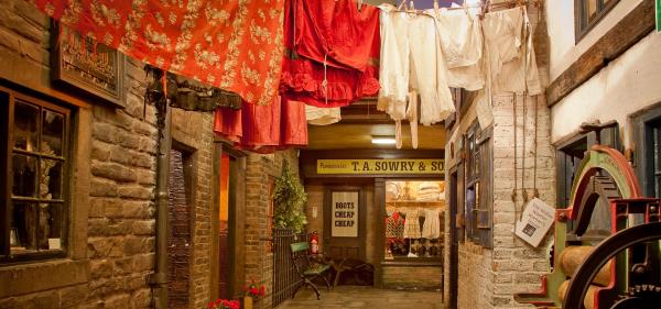 A colour photograph of a recreated Victorian backstreet (Abbey Fold at Abbey House Museum), featuring different dwellings. Anti-clockwise from the right is the widow washerwoman's house, with her mangle. Next is the artisan's cottage with whitewashed walls. In the centre is the pawnbrokers shop with the pawnbrokers house top left, leading down to the mourning warehouse window in the bottom left corner.