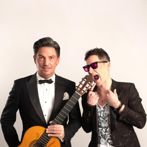 A man in a classic tuxedo with a slight coiffed haircut holds an acoustic guitar alongside a man in a patterned suit jacket, vest and purple sunglasses who is mimicking wildly eating the guitar.