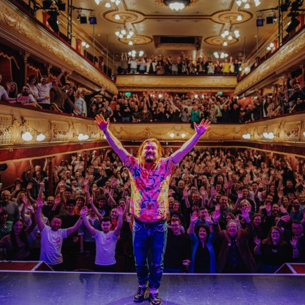 A man poses on stage with his arms in the air, in front of a packed crowd mimicking the same gesture.