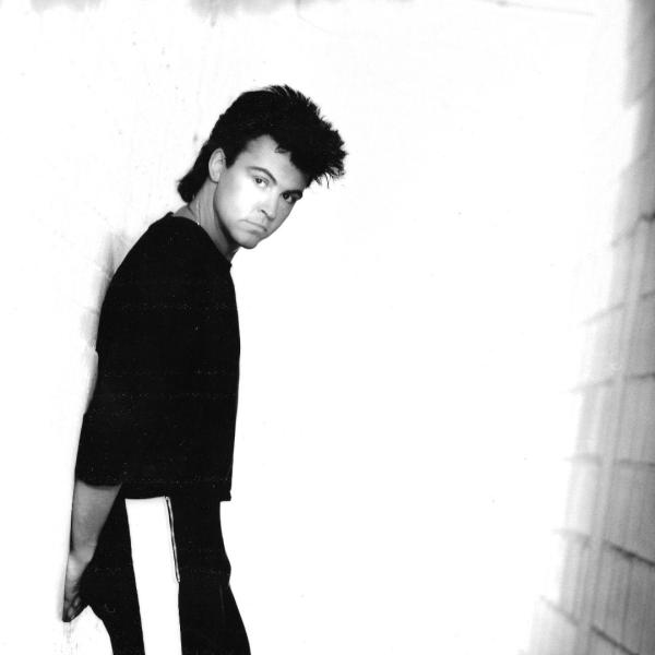 Black and white photo of Paul Young leaning against a wall, wearing a long sleeve black t-shirt.