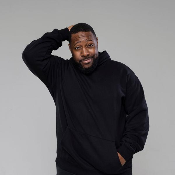 Aurie Styla smirks, with one hand held behind his head, the other in the pocket of his black hoodie, pictured against a grey backdrop.