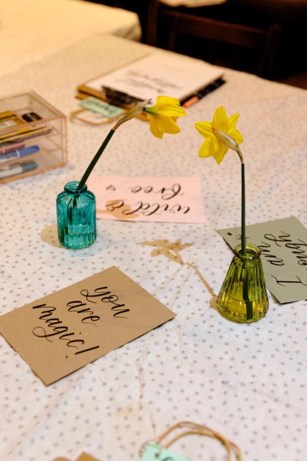 Image shows calligraphy samples of affirmations on various coloured cards on a table top. Daffodils sit in vases and a calligraphy pen can be seen in the background.