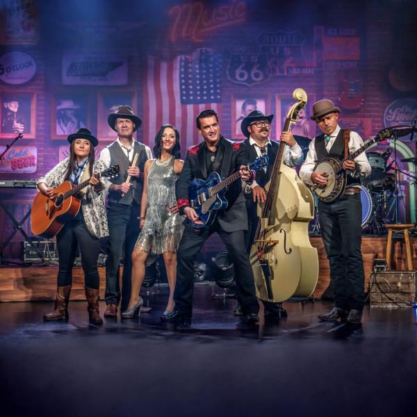 The members of A Country Night in Nashville stand together on stage in period costume, each holding their guitar or instrument. American memorabilia, photographs and flags are visible in the background.