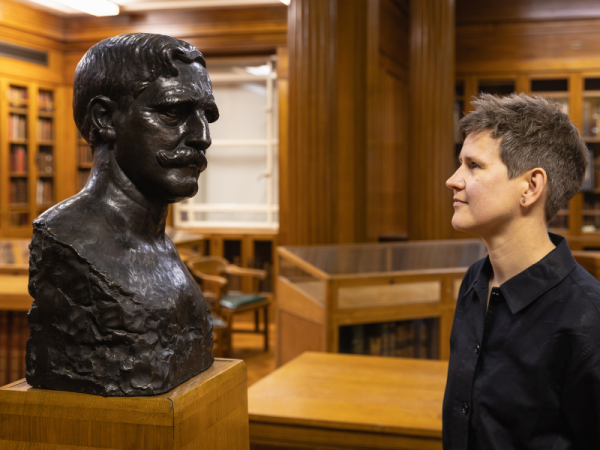 A person with short brown hair and wearing a black shirt looks at a bronze bust of a man with a moustache on a wooden plinth. They are in a wood-panelled room.