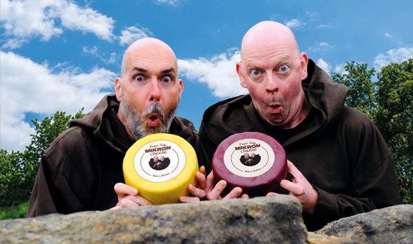 Two bald monks peer over a dry stone wall in Wensleydale looking shocked or excited while holding large cheeses
