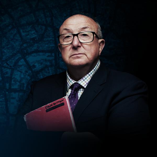 Colin Sutton in a suit and glasses with arms folded around a red notebook.