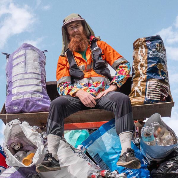 A man with a long ginger beard, a floral shirt and orange high-vis jacket sits on top of a small mountain of bin bags containing cans and plastic bottles in front of a cloudy sky.