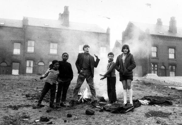 Six children stand around a small fire on a rubble-strewn urban street, with rows of terraced houses behind them and smoke rising in the background.