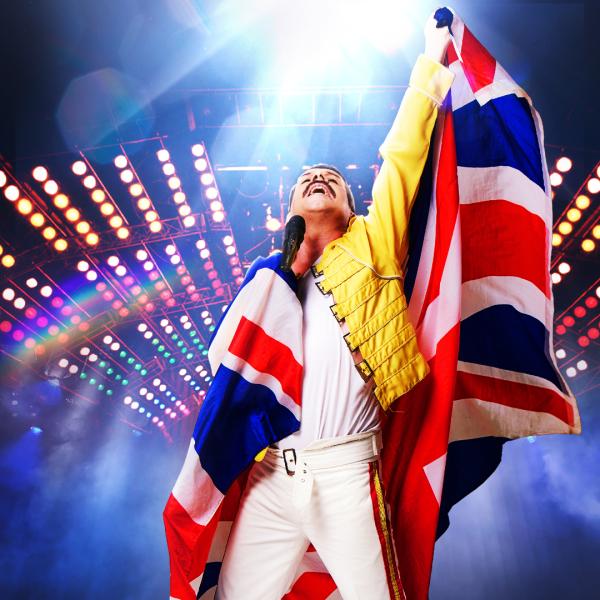 A Freddie Mercury tribute singer in a yellow jacket, white trousers and a Union flag draped over his shoulders raises a fist, in front of rows of spotlights. 